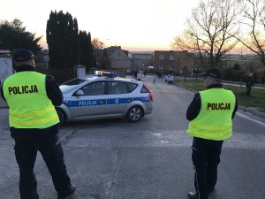 Na pierwszym planie zdjęcia stoi dwóch umundurowanych policjantów, przed nimi oznakowany radiowóz. w oddali widać uczestników procesji.