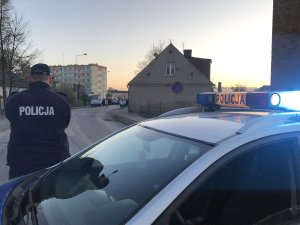 Na pierwszym planie zdjęcia widać oznakowany radiowóz, przed nim umundurowanego policjanta obserwującego idących ulicą uczestników procesji.