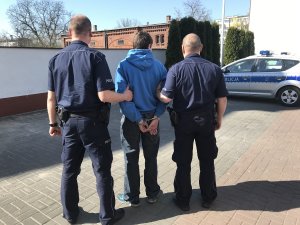 Zdjęcie przedstawia dwóch umundurowanych policjantów prowadzących w kajdankach zatrzymanego. w tle zdjęcia. po prawej stronie widać oznakowany radiowóz.