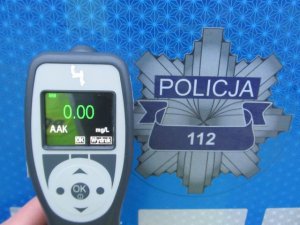 Zdjęcie przedstawia gwiazdę policyjną koloru szarego z napisem Policja oraz numerem alarmowym 112. Gwiazda umiejscowiona jest na tle koloru niebieskiego. Gwiazda stanowi tłu fotografii. Na pierwszym planie, po lewej stronie znajduje się urządzenie do badania stanu trzeźwości