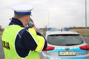 Zdjęcie przedstawia na pierwszym planie, po lewej stronie, umundurowanego policjanta ruchu drogowego mierzącego prędkość pojazdów urządzeniem do pomiaru prędkości. Po prawej stronie zdjęcia, w tle widać oznakowany policyjny radiowóz.