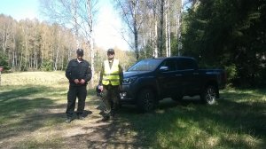 Zdjęcie przedstawia umundurowanego policjanta stojącego w lesie wspólnie z przedstawicielem straży leśnej. Na drugim planie zdjęcia widać nieoznakowany pojazd straży leśnej