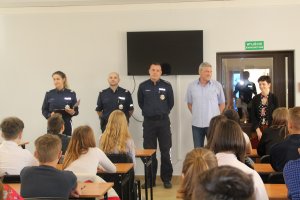 Fotografia przedstawia siedzących w ławkach uczestników konkursu. a na drugim planie stoją umundurowani policjanci omawiający uczestnikom założenia konkursu.