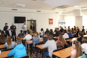 Fotografia przedstawia siedzących w ławkach uczestników konkursu. a na drugim planie stoją umundurowani policjanci omawiający uczestnikom założenia konkursu.