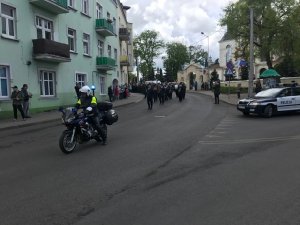 na zdjęciu policjant na motorze w tle z prawej strony radiowóz a w tle idą ludzie w pochodzie