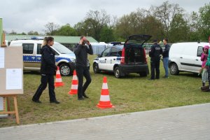 Fotografia przedstawia umundurowaną policjantkę nadzorującą uczestnika festynu w Minikowie podczas pokonywania toru przeszkód z założonymi alkogoglami. na drugim tle widać umundurowanego policjanta rozmawiającego z uczestniczką targów. W tle również znajdują się oznakowane radiowozy.