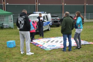 Fotografia przedstawia umundurowaną policjantkę prowadzącą grę edukacyjną dla dzieci obok niej dwoje dzieci. na pierwszym planie stoją trzy osoby dorosłe, kobietę oraz dwóch mężczyzn, odwróconych plecami do zdjęcia. w tle oznakowany radiowóz oraz namiot wystawowy koloru zielonego.