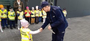 Zdjęcie przedstawia umundurowanego policjanta wręczającego dziewczynce odblask. w tle widać grupę przedszkolaków ubranych w kamizelki odblaskowe.