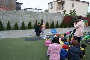 Fotografia przedstawia umundurowaną policjantkę uczącą stojącego koło niej chłopca właściwego korzystania z przejścia dla pieszych. na pierwszym planie widać przyglądające się dzieci oraz ich opiekunkę.