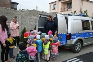 Zdjęcie przedstawia umundurowaną policjantkę prezentującą przedszkolakom oznakowany radiowóz. Na fotografii widać tez grupę przedszkolaków oraz dwie opiekunki dzieci.