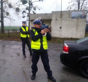 Na pierwszym planie widać umundurowanego policjanta ruchu drogowego wykonującego pomiar prędkości, w tle stoi drugi umundurowany policjant drogówki.