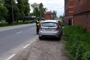 Zdjęcie przedstawia umundurowanego policjanta ruchu drogowego prowadzącego kontrolę stojącego przed nim auta. Obok pojazdu stoją też dwie osoby.