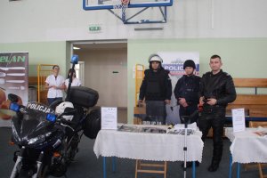 Zdjęcie przedstawia umundurowanych policjantów biorących udział w targach pracy. w tle widać baner Komendy Powiatowej Policji w Nakle nad Notecią. na pierwszym planie zdjecia przed policjantami po lewej stronie fotografii stoi policyjny motocykl. Jeden z policjantów ubrany ma na sobie kombinezon do jazdy motocyklem, a dwoje ubranie specjalistyczne wykorzystywane podczas zabezpieczeń rożnego rodzaju imprez sportowych i masowych.