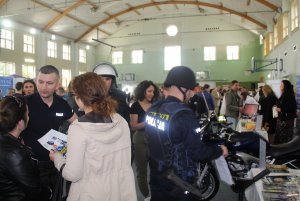 Zdjęcie przedstawia umundurowanych policjantów rozmawiających z uczestnikami targów. dwóch z policjantów ubranych w ubranie specjalistyczne wykorzystywane podczas zabezpieczeń rożnego rodzaju imprez sportowych i masowych. Trzeci w kombinezon motocyklowy.