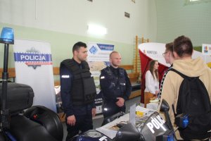 Zdjęcie przedstawia umundurowanych policjantów rozmawiających z uczestnikami targów. W tle widać baner Komendy Powiatowej Policji w Nakle nad Notecią. na pierwszym planie zdjęcia przed policjantami po lewej stronie fotografii widoczny element policyjnego motocykla. Policjanci ubrani w ubrania specjalistyczne wykorzystywane podczas zabezpieczeń rożnego rodzaju imprez sportowych i masowych.