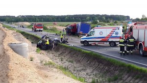 Fotografia przedstawia miejsce wypadku drogowego. po prawej stronie widoczne wozy straży pożarnej, strażaków oraz karetkę pogotowia. w tle rozbity pojazd cieżarowy. po lewej stronie zdjęcia zniszczony pojazd osobowy i stojący obok niego policjanci ubrani w żółte kamizelki odblaskowe.