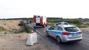 Fotografia przedstawia miejsce wypadku drogowego. na pierwszym planie oznakowany policyjny radiowóz, w tle wozy straży pożarnej i rozbite auta.