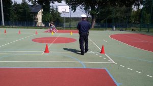 Zdjęcie przedstawia umundurowanego policjanta ruchu drogowego stojącego na płycie boiska szkolnego i prowadzącego egzamin praktyczny na kartę rowerową. przed policjantem w tle widać dziewczynkę jadącą rowerem.