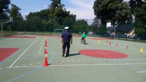 Zdjęcie przedstawia umundurowanego policjanta ruchu drogowego stojącego na płycie boiska szkolnego i prowadzącego egzamin praktyczny na kartę rowerową. przed policjantem w tle widać chłopca jadącego rowerem.