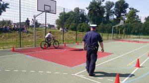 Zdjęcie przedstawia umundurowanego policjanta ruchu drogowego stojącego na płycie boiska szkolnego i prowadzącego egzamin praktyczny na kartę rowerową. po lewej stronie policjanta widać chłopca jadącego rowerem. w tle, za płotem boiska widać uczniów siedzących na skarpie i obserwujących prowadzony egzamin.