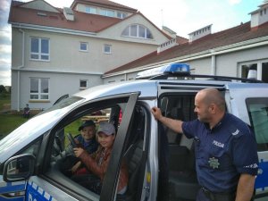 Zdjęcie przedstawia na pierwszym planie umundurowanego policjanta trzymającego otwarte drzwi oznakowanego radiowozu. we wnętrzu pojazdu siedzi dwoje dzieci.