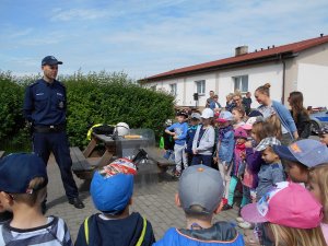 Zdjęcie przedstawia umundurowanego policjanta stojącego przodem do fotografii i prowadzącego zajęcia dla stojących przed nim dziećmi.