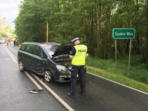 Zdjęcie przedstawia miejsce wypadku drogowego. na pierwszym planie stoi umundurowany policjant ruchu drogowego w żółtej kamizelce. po jego prawej stronie tablica z nazwą miejscowości - Szubin Wieś. przed policjantem rozbity pojazd. w tle widać stojące pojazdy między innymi karetkę oraz strażaków.