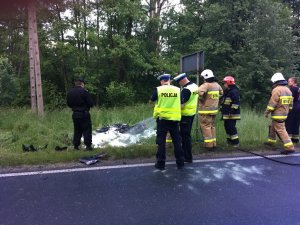 Zdjęcie przedstawia umundurowanych funkcjonariuszy policji i straży pożarnej stojących w rejonie drogi. przed nimi na trawie wrak motoroweru.