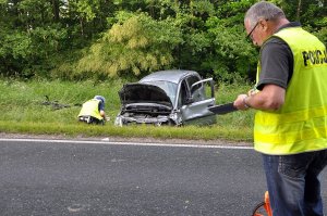 Zdjęcie przedstawia miejsce wypadku drogowego. na pierwszym planie stoi nieumundurowany policjant pionu kryminalnego ubrany w żółtą kamizelkę. Na wprost w oddali rozbity samochód przy którym oględziny wykonuje policjant ruchu drogowego.