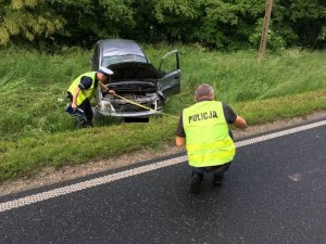 Zdjęcie przedstawia miejsce wypadku drogowego. na pierwszym planie stoi nieumundurowany policjant pionu kryminalnego ubrany w żółtą kamizelkę. Na wprost w oddali rozbity samochód przy którym oględziny wykonuje policjant ruchu drogowego.