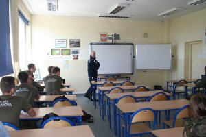 Zdjęcie przedstawia umundurowanego policjanta prowadzącego zajęcia dla siedzących przed nim w klasie uczniów.