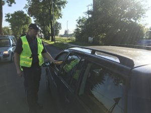 Zdjęcie przedstawia umundurowanego policjanta, dodatkowo ubranego w żółtą kamizelkę. Policjant prowadzi kontrolę trzeźwości kierowcy przejeżdżającego auta.