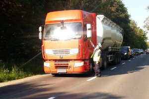 Zdjęcie przedstawia umundurowanego policjanta, dodatkowo ubranego w żółtą kamizelkę. Policjant prowadzi kontrolę trzeźwości kierowcy przejeżdżającego auta.