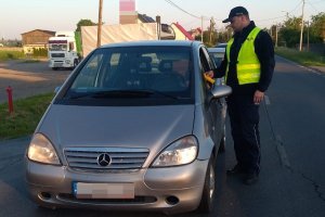 Zdjęcie przedstawia umundurowanego policjanta, dodatkowo ubranego w żółtą kamizelkę. Policjant prowadzi kontrolę trzeźwości kierowcy przejeżdżającego auta.