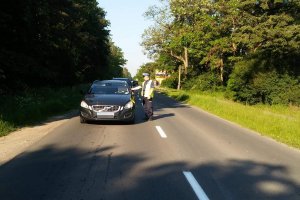 Zdjęcie przedstawia umundurowanego policjanta, dodatkowo ubranego w żółtą kamizelkę. Policjant prowadzi kontrolę trzeźwości kierowcy przejeżdżającego auta.