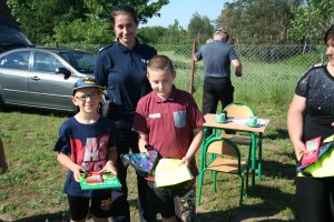 Zdjęcie przedstawia stojącą umundurowaną policjantkę, a przed nią dwoje dzieci. w tle oznakowany radiowóz.