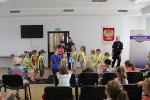 Zdjęcie przedstawia umundurowanych policjantów - kobietę i dwóch mężczyzn prowadzących zajęcia na temat bezpieczeństwa w ruchu drogowym dla siedzących na sali dzieci. część dzieci ubrane ma kamizelki odblaskowe i ćwiczy pod okiem policjantów poznane przepisy drogowe na mini miasteczku ruchu drogowego.