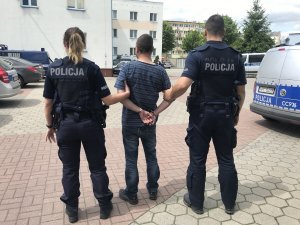 Zdjęcie przedstawia osobę zatrzymaną prowadzoną przez umundurowanych policjantów przez plac komendy. Osoba ma założone kajdanki na trzymane z tyłu dłonie.