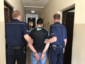 Zdjęcie przedstawia zatrzymanego prowadzonego przez umundurowanych policjantów. Zatrzymany na trzymanych z tyłu dłoniach założone ma kajdanki.