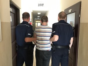 Zdjęcie przedstawia zatrzymanego prowadzonego przez umundurowanych policjantów.