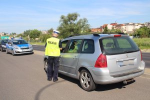 Zdjęcie przedstawia umundurowanego policjanta ruchu drogowego kontrolującego stojący przed nim samochód osobowy. Policjant ma ubraną tez żółtą kamizelkę . w tle widac oznakowany policyjny radiowóz.