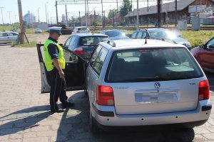 Zdjecie przedstawia umundurowanego policjanta ruchu drogowego kontrolującego pojazd osobowy. w tle widać inne pojazdy oraz torowisko.