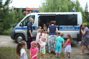 Zdjęcie przestawia dzieci stojące na pierwszym planie. w tle policyjny oznakowany radiowóz obok niego umundurowany policjant.