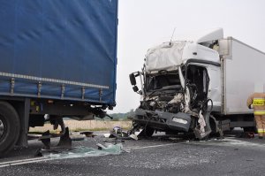 Zdjęcie przedstawia naczepę pojazdu ciężarowego koloru niebieskiego. za nią rozbity ciężarowy mercedes.