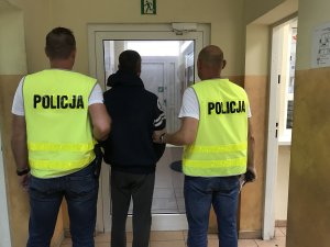 Zdjęcie przedstawia nieumundurowanych policjantów, ubranych w żółte kamizelki z napisem policja, prowadzących zatrzymanego mężczyznę.