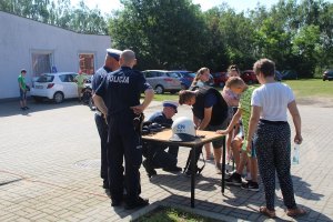 Zdjęcie przedstawia chłopca, którego policjant ubiera w specjalistyczne ubranie. Obok stoją inne dzieci i dwóch umundurowanych policjantów.