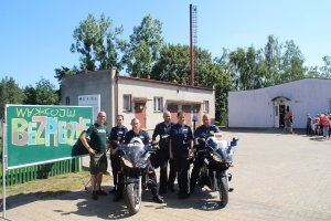 Zdjęcie przedstawia umundurowanych policjantów: dwie kobiety, trzech mężczyzn. Dwóch policjantów siedzi na służbowych motocyklach. Obok funkcjonariuszy stoi pracownik szkoły. Wszyscy stoją przy tablicy, na której umieszczony jest kolorowy napis drukowanymi literami BEZPIECZNE WAKACJE