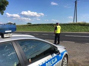 Zdjęcie przedstawia na pierwszym planie oznakowany radiowóz. w tle widać umundurowanego policjanta prowadzącego pomiar prędkości urządzeniem iskra.