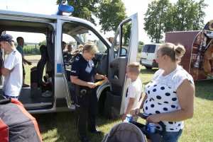 zdjęcie przedstawia na pierwszym planie kobietę z wózkiem. W tle radiowóz oznakowany policji, przed nim stoi umundurowana policjantka, która prezentuje sprzęt będący na wyposażeniu policji. obok niej uczestnicy festynu.