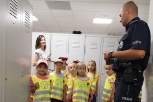 Zdjęcie przedstawia dzieci wraz z opiekunkami i umundurowanym policjantem zwiedzające szatnię w komisariacie.